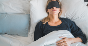 Woman lying in bed with a black sleep mask over her eyes, tucked under a white blanket.
