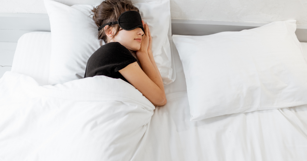 Woman resting in bed with a black sleep mask over her eyes, tucked under white sheets and pillows.