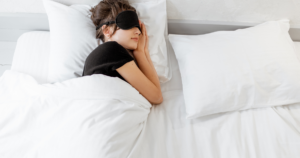 Woman resting in bed with a black sleep mask over her eyes, tucked under white sheets and pillows.