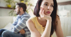 A thoughtful woman rests her chin on her hand while a man sits away in the background, implying relationship strain in a cafe-like living space.
