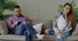 Man in a red plaid shirt sits on a beige couch reading a tablet while a woman with long hair sits nearby, clutching a white pillow and looking down in a living room with plants and a white brick wall.