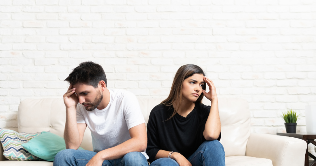 Couple sitting on a white couch against a white brick wall, both looking stressed and touching their heads in worry.