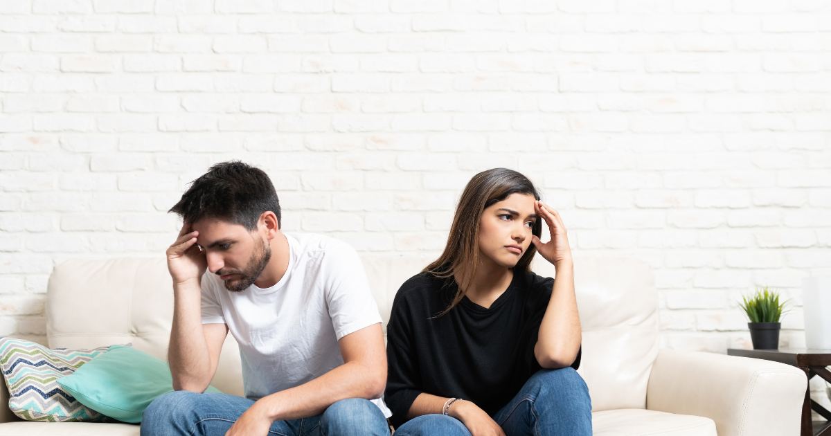 Couple sitting on a white couch against a white brick wall, both looking stressed and touching their heads in worry.