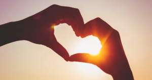 Hands form a heart shape around the sun as it shines through at sunset/ sunrise.