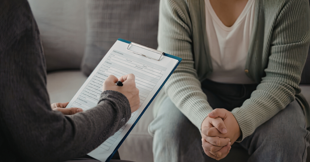 Therapist writes on a clipboard while client sits on a sofa with hands clasped, during a session.