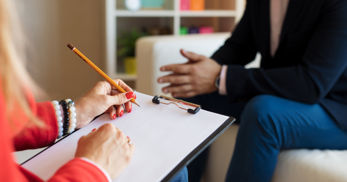 Two people in a counseling-like setting: one person writes on a clipboard with a pencil while the other speaks and gestures hand motions in the background.