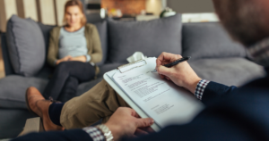Therapist taking notes on a clipboard while a patient sits on a couch in a living room