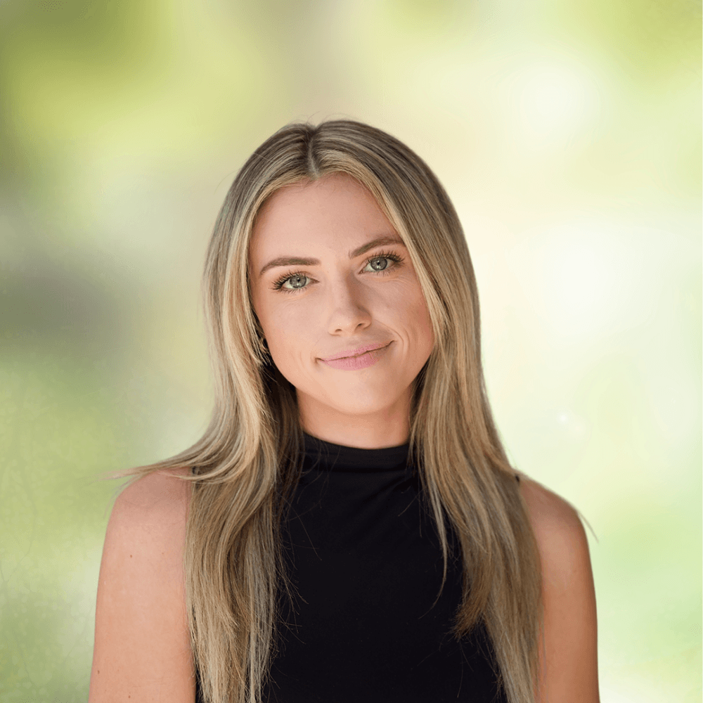 Close-up portrait of a smiling blonde woman wearing a black sleeveless top, outdoors with a soft green background.