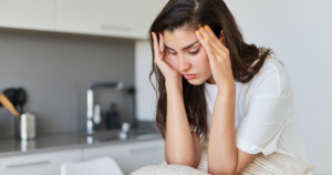 Woman in a white top sitting in a modern kitchen, rubbing her temples with a worried expression.