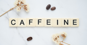 Scrabble-style tiles spell'CAFFEINE' on a light marble surface, with a few coffee beans and dried flowers nearby
