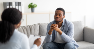Man wearing a blue shirt sits on a gray sofa, hands clasped, during a counseling session with a therapist taking notes nearby (focus on conversation).]