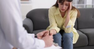 女の人がソファに座り、手で頭を押さえて困惑した表情。カウンセリングを受けている場面。治療者がメモを取っている。