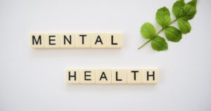 Scrabble tiles spell'MENTAL HEALTH' on a white background with a green leafy branch to the right, conveying mental health awareness.