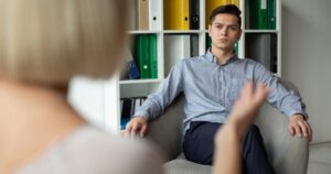 Man in a light button-down shirt sits in an armchair, listening during a one-on-one counseling session.