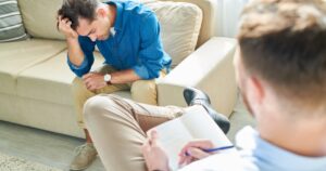 Man sits on a sofa, head in hand, during a therapy session with a clinician taking notes nearby in a living room setting.