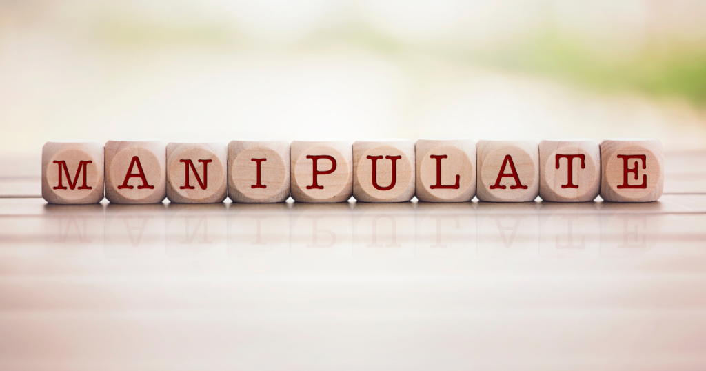 Wooden letter blocks spell MANIPULATE on a reflective surface with a soft, blurred background