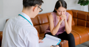 - Shine Mental Health Clinic consultation: a man clinician with a stethoscope reviews notes on a clipboard as a worried woman sits on a brown leather sofa, hands clasped.