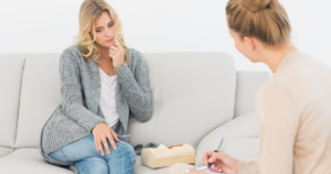 Woman sitting on a light sofa in a gray cardigan, touching her face, as a therapist nearby takes notes and a tissue box sits on the table between them.