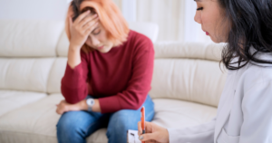 Distressed woman with red hair sits on a sofa, hand on her forehead, while a clinician in a white coat speaks to her in a living room setting.