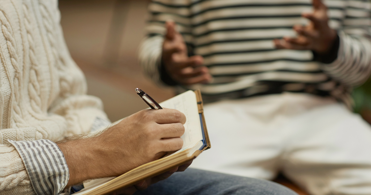 Person writes in a notebook with a pen while another person gestures in the background.