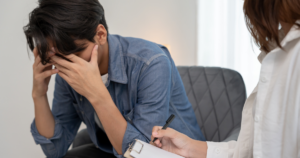 Man sits with his head in his hands during a therapy session as a clinician takes notes on a clipboard when speaking to him?