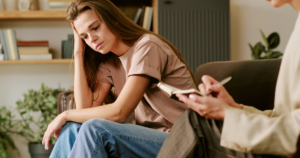 Woman sits on a couch with her head resting on her hand, appearing sad during a therapy session as a counselor takes notes nearby.