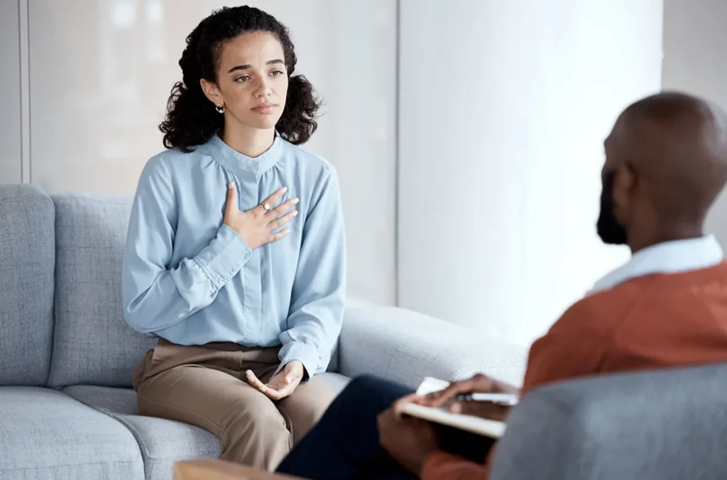 Woman in a light blue blouse sits on a sofa, hand over heart, speaking with a therapist in a counseling session.