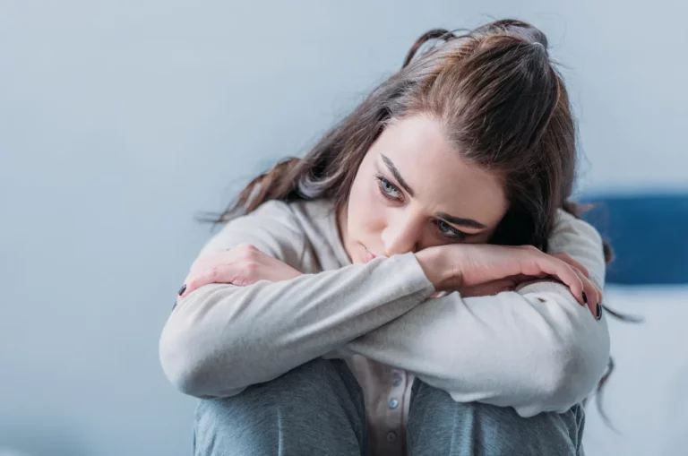 Young woman sits with her arms wrapped around her knees, resting her head on her forearms, looking sad and introspective.