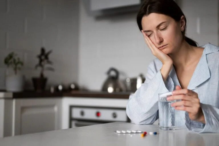 Tired woman sits at a kitchen table with a glass of water, a blister pack of pills on the table, and a hand resting on her cheek.