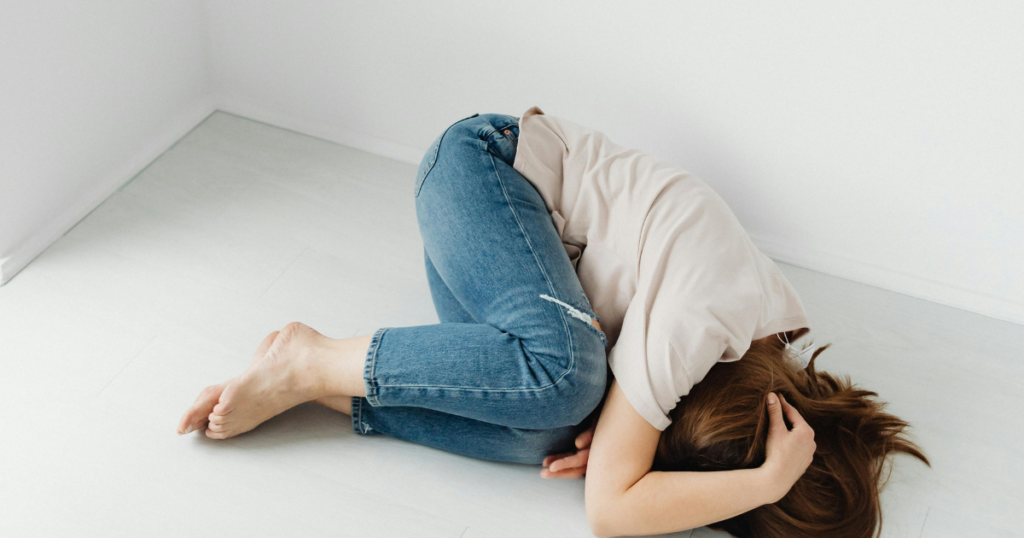 feeling-of-impending-doom-body-triggers-image-2 - Shine Mental Health Person curled on the floor in a white room, knees drawn up, wearing jeans and a beige shirt.