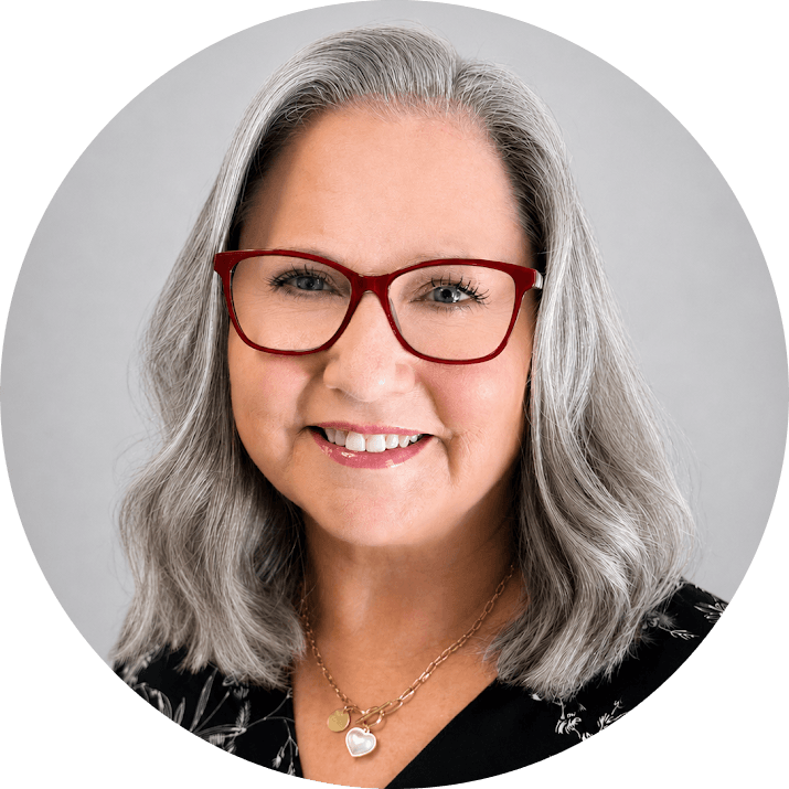 Catherine 1 - Shine Mental Health Smiling woman with shoulder-length gray hair and red glasses wearing a black patterned top and a heart pendant necklace, neutral gray background.