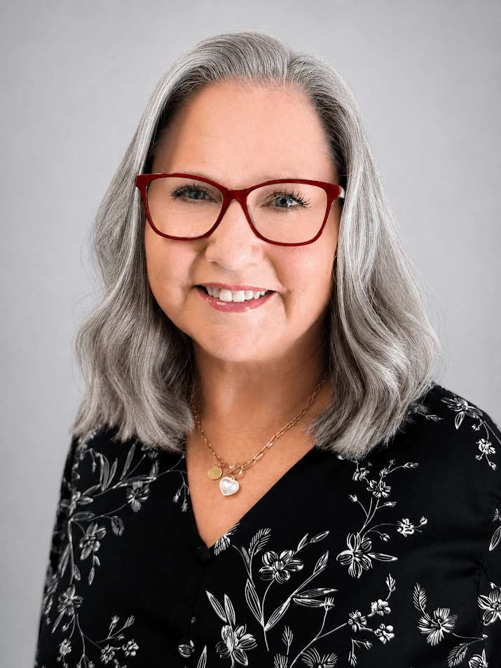 Smiling gray-haired woman with red-framed glasses wearing a black floral blouse and a heart pendant necklace.