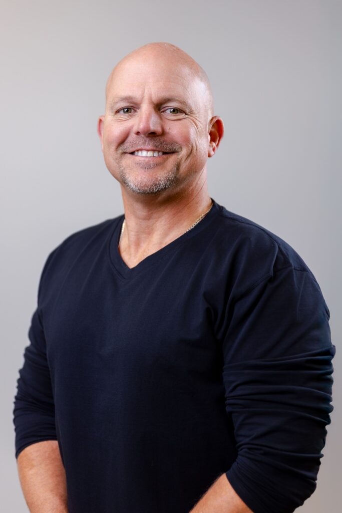 Smiling bald man in a navy crewneck sweater, posing for a portrait against a light gray background.