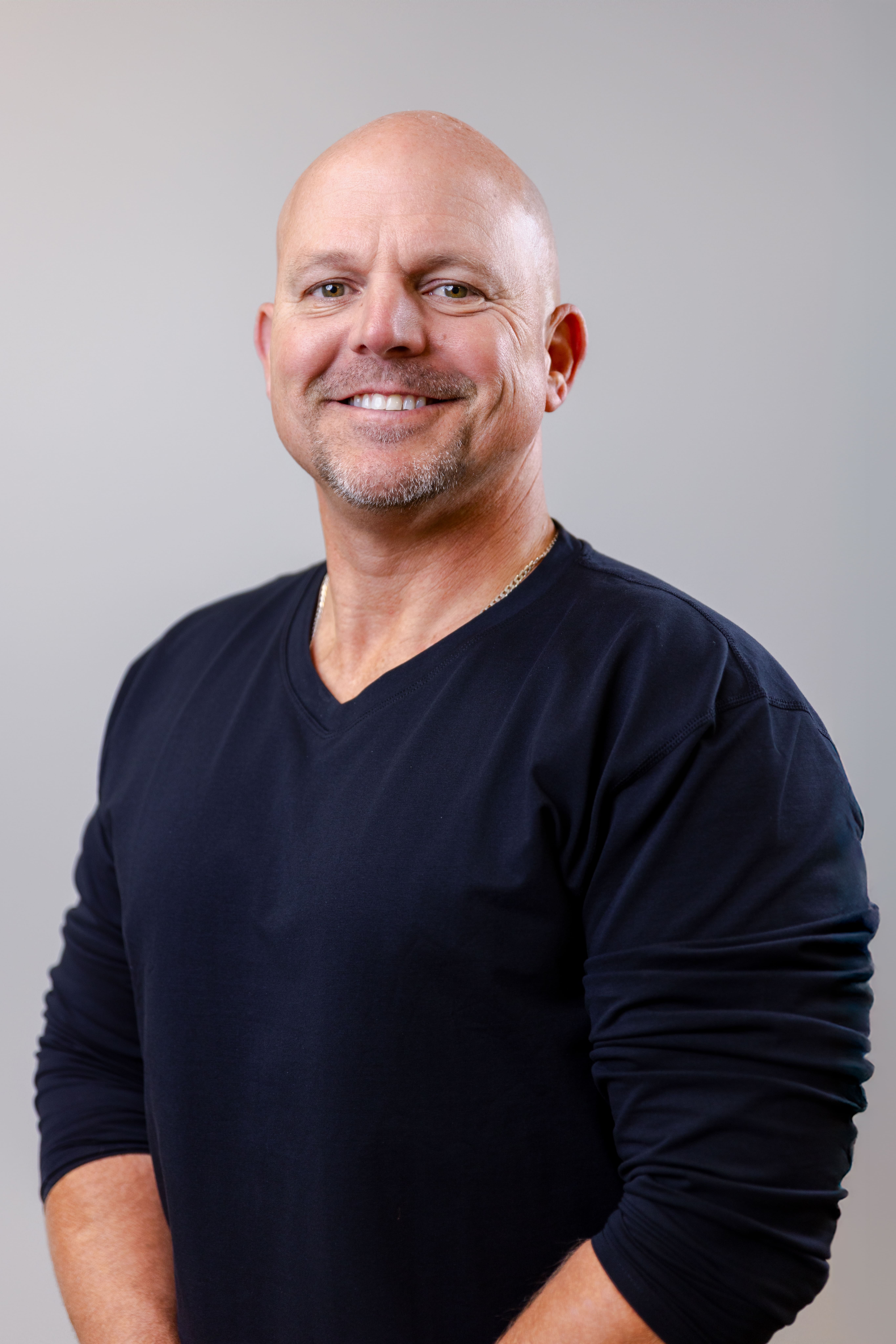 Smiling bald man in a navy crewneck sweater, posing for a portrait against a light gray background.