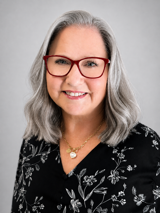 Smiling woman with shoulder-length gray hair and red glasses, wearing a black floral blouse and gold heart necklace.