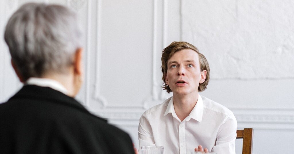 Young man in a white shirt speaks during a meeting, facing an older person with gray hair.