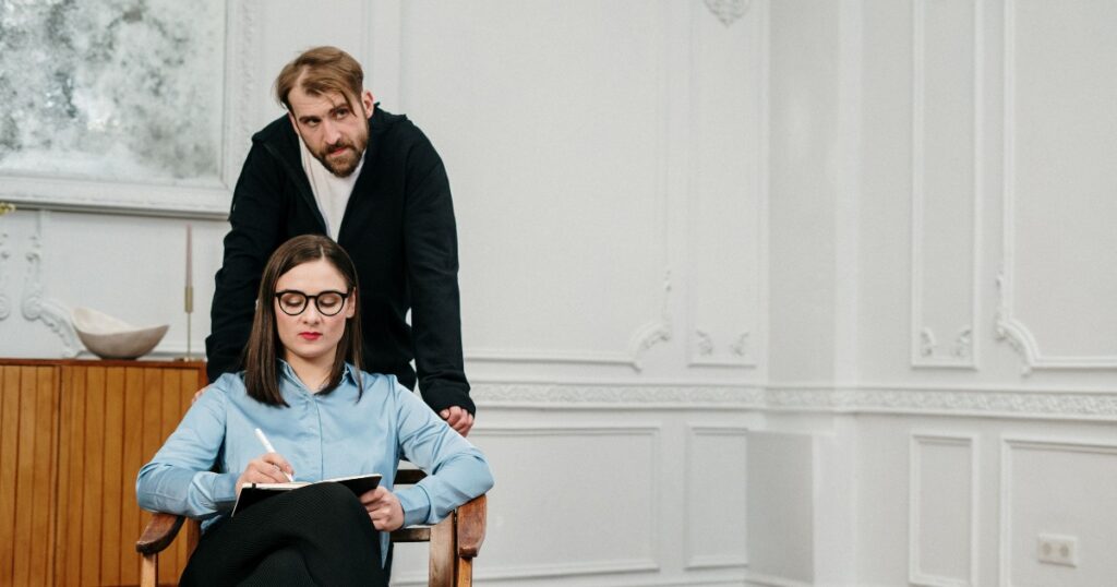 Man stands behind a seated woman who writes in a notebook on a wooden chair in a bright, white-paneled room.