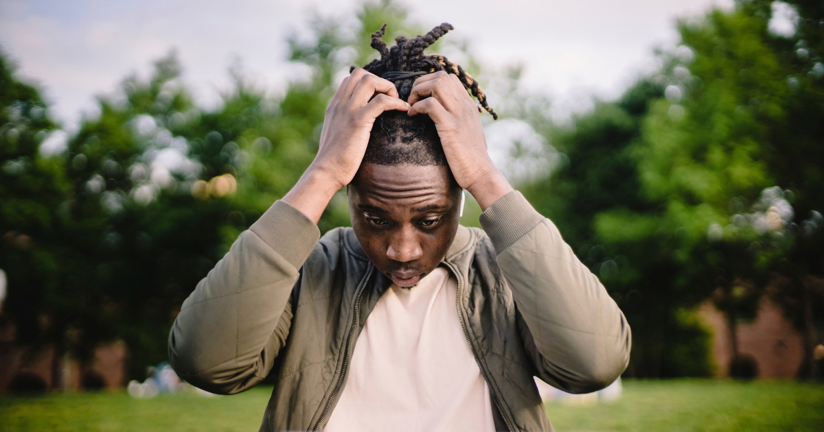 Young man in a beige jacket stands in a park, hands on his head, looking stressed or overwhelmed.