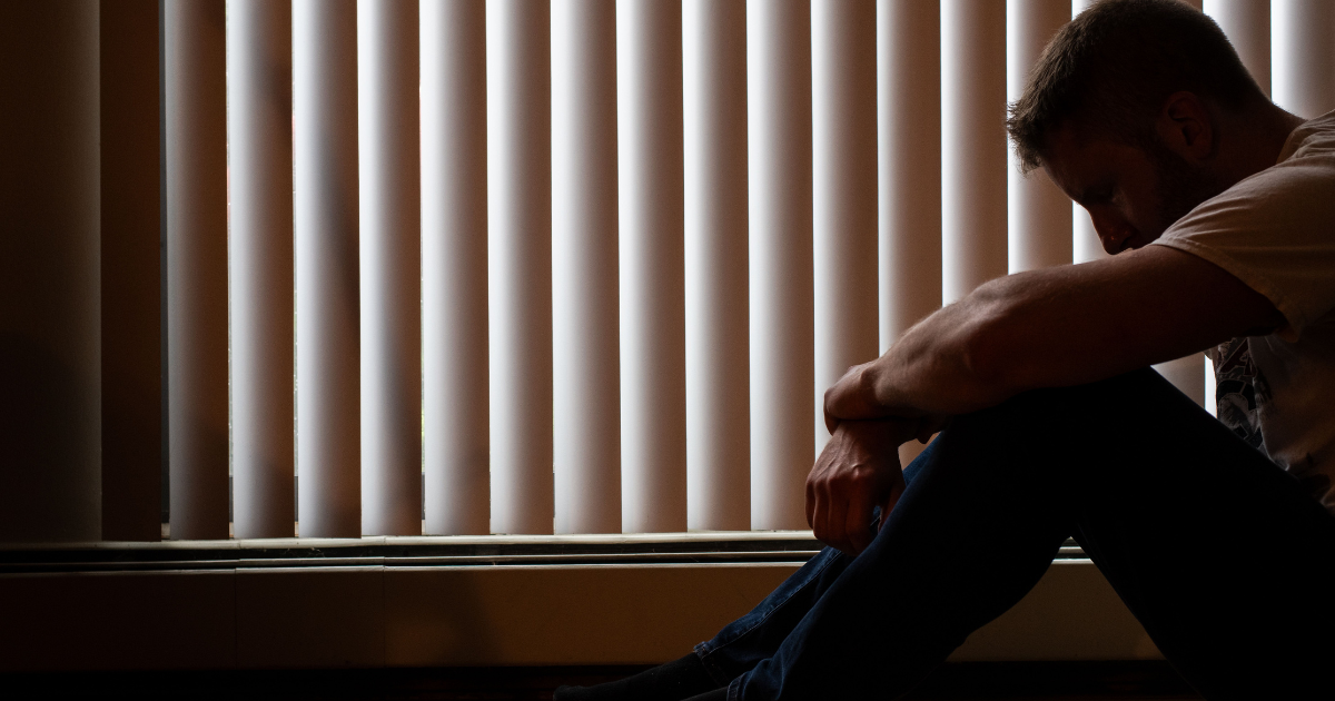Man sits on the floor beside vertical blinds, head bowed in a dimly lit room, conveying a somber mood.