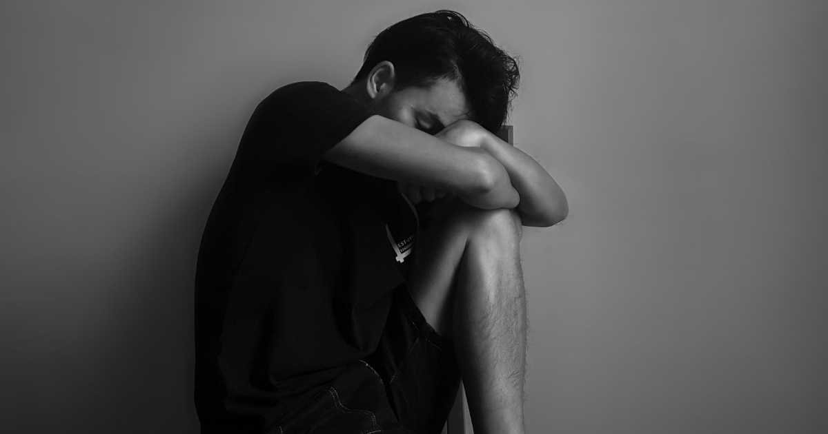 Young man sitting against a wall, arms wrapped around his knees, head resting on his arms in sadness/withdrawal.