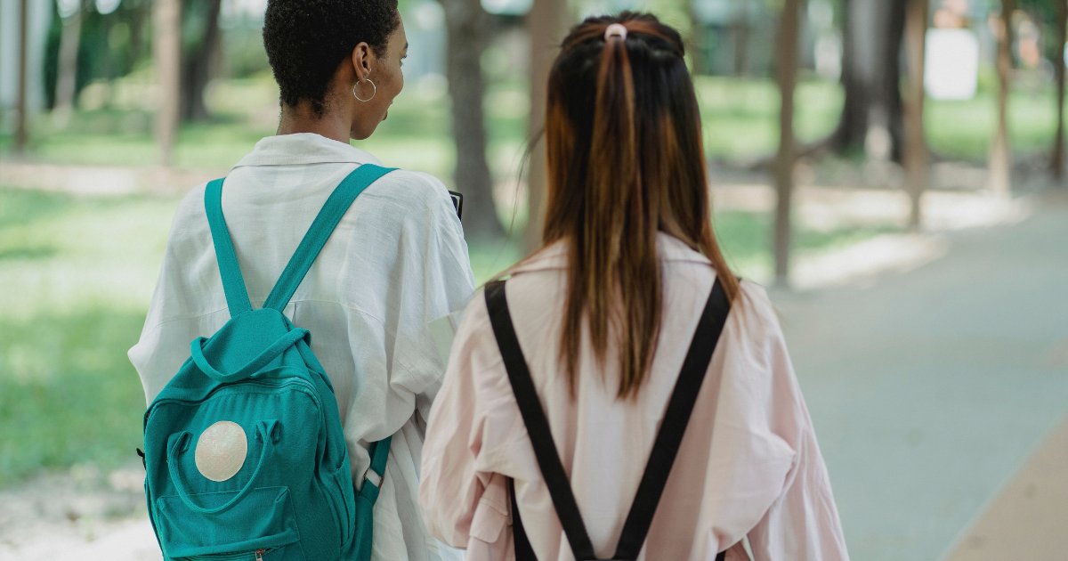 Two people walk side by side in a park, seen from behind; one with a teal backpack and white shirt, the other with pale pink top and black suspenders.