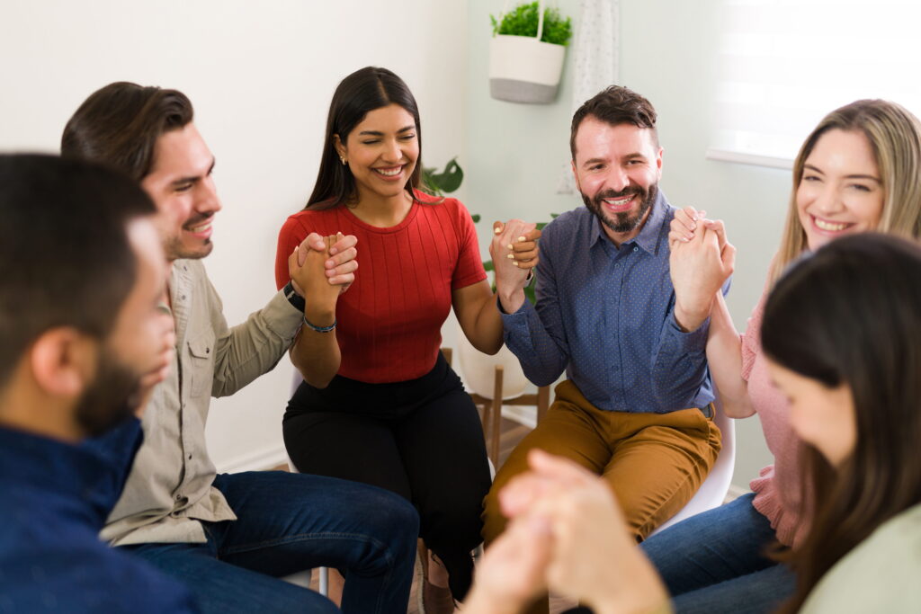 Group of friends or coworkers sitting in a circle, hands joined, smiling in a bright room. This conveys unity and camaraderie during a meetup.