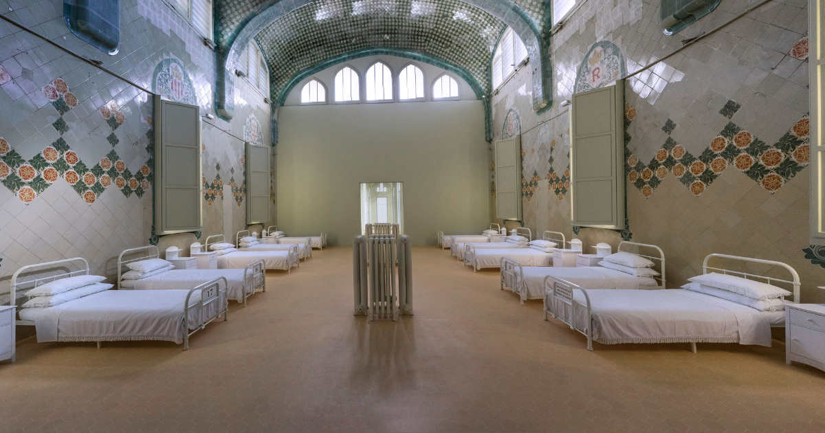 A long ward with rows of metal-framed beds along tiled walls, arched windows, and decorative ceramic tiles on the walls and ceiling. Empty hospital dormitory room.