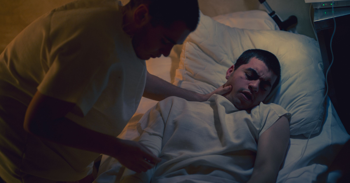 Caregiver checks the patient’s neck while the man lies in a hospital bed at night under a blanket.