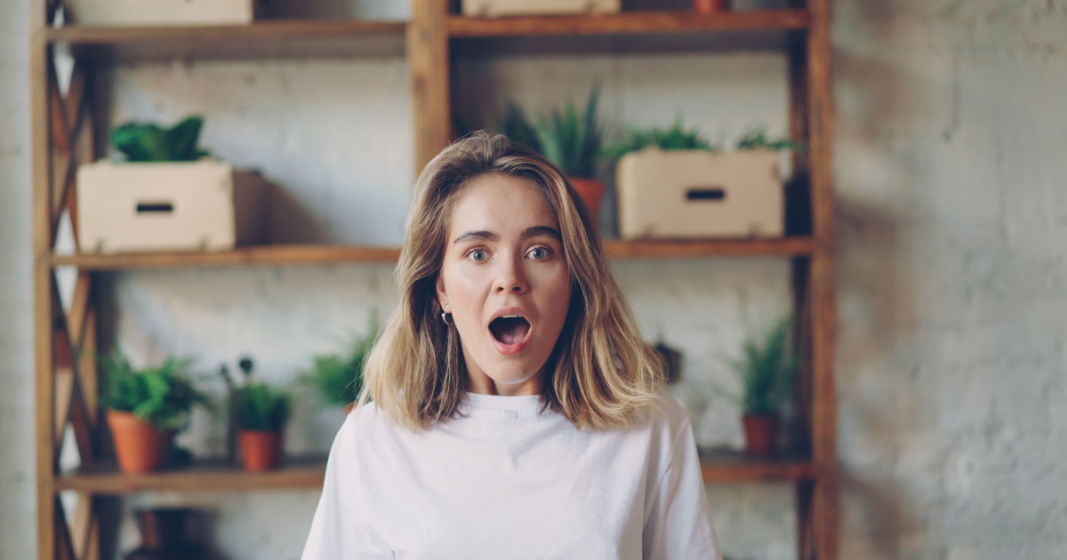 Surprised woman in a white shirt with shoulder-length blonde hair, standing in front of wooden shelves with potted plants.