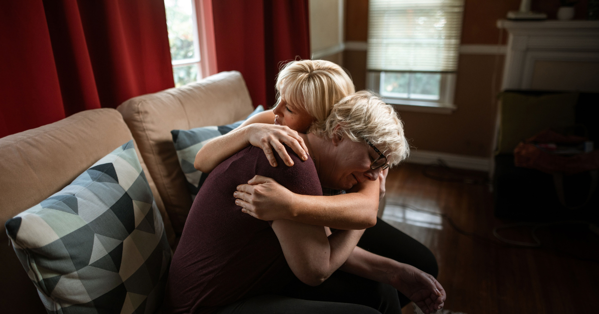 Two people share a warm hug on a beige sofa in a cozy living room with red curtains.
