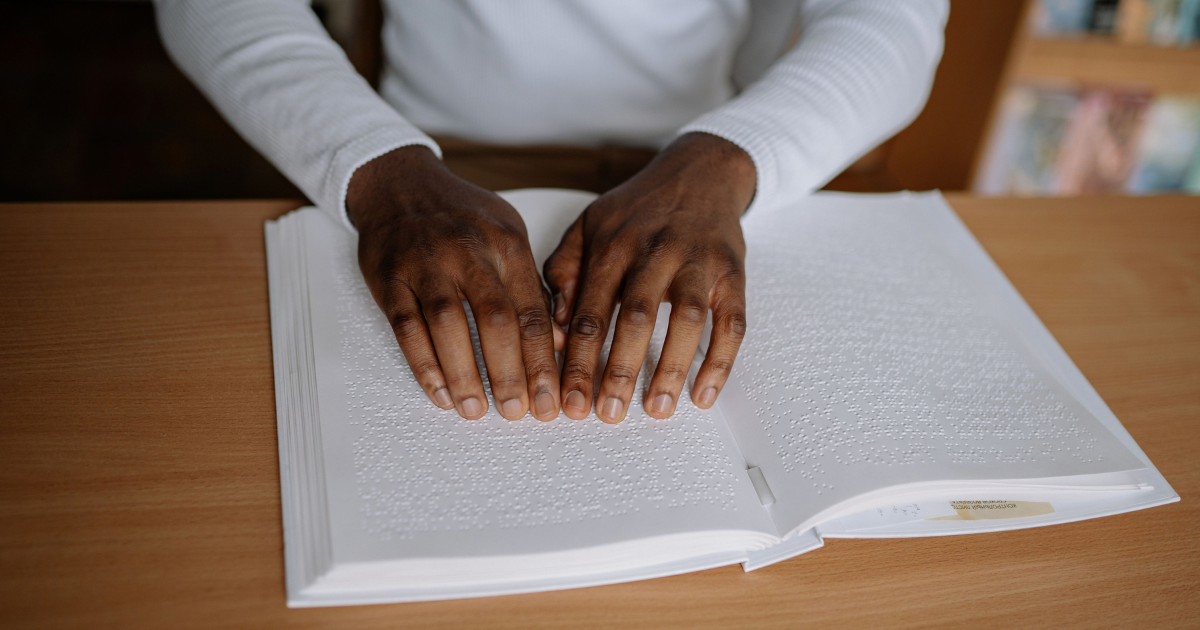 Person reading a Braille book with both hands on tactile pages at a wooden table.