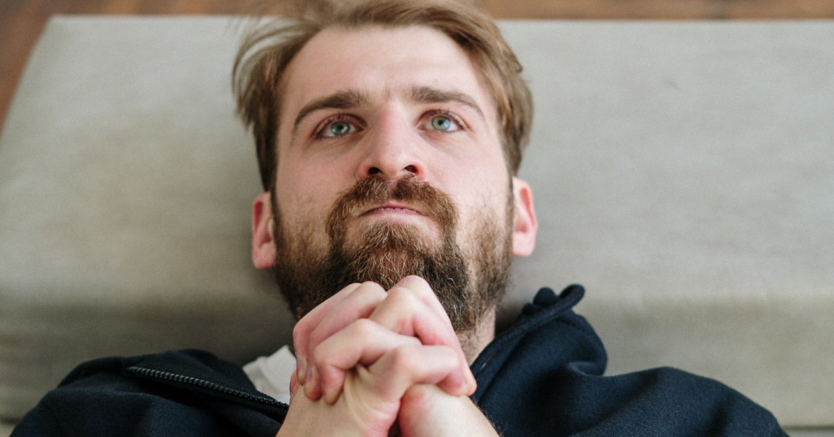 Bearded man lying on his back, hands clasped under his chin, looking upward