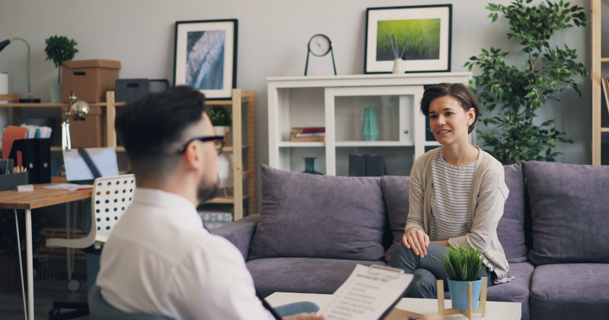 Woman seated on a purple sofa converses with a man in a modern, plant-filled office setting with shelves and framed art behind them, suggesting a casual meeting.