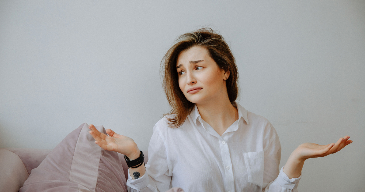 A young woman in a white shirt shrugs with a puzzled expression, palms up, seated on a pink couch against a light gray wall.